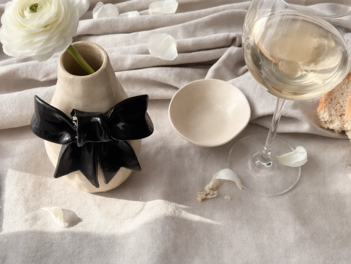A cream ceramic bud vase with a black bow holding a white ranunculus on rumpled linen, beside a small ceramic dish and a glass of white wine